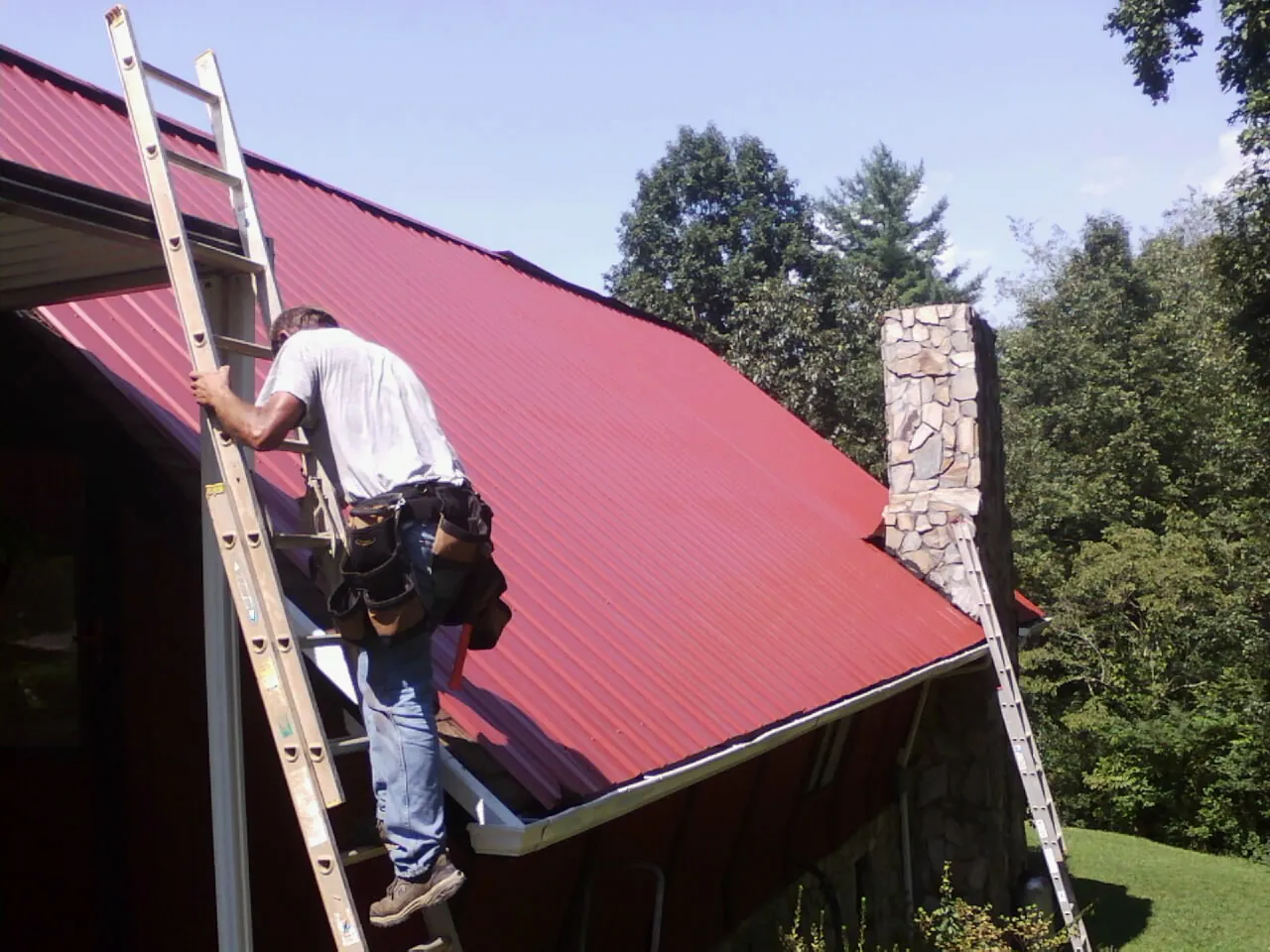 Roofer on a ladder beside a red metal roof and stone chimney — Soffit & Fascia Repair by Roofing Brothers in Story City